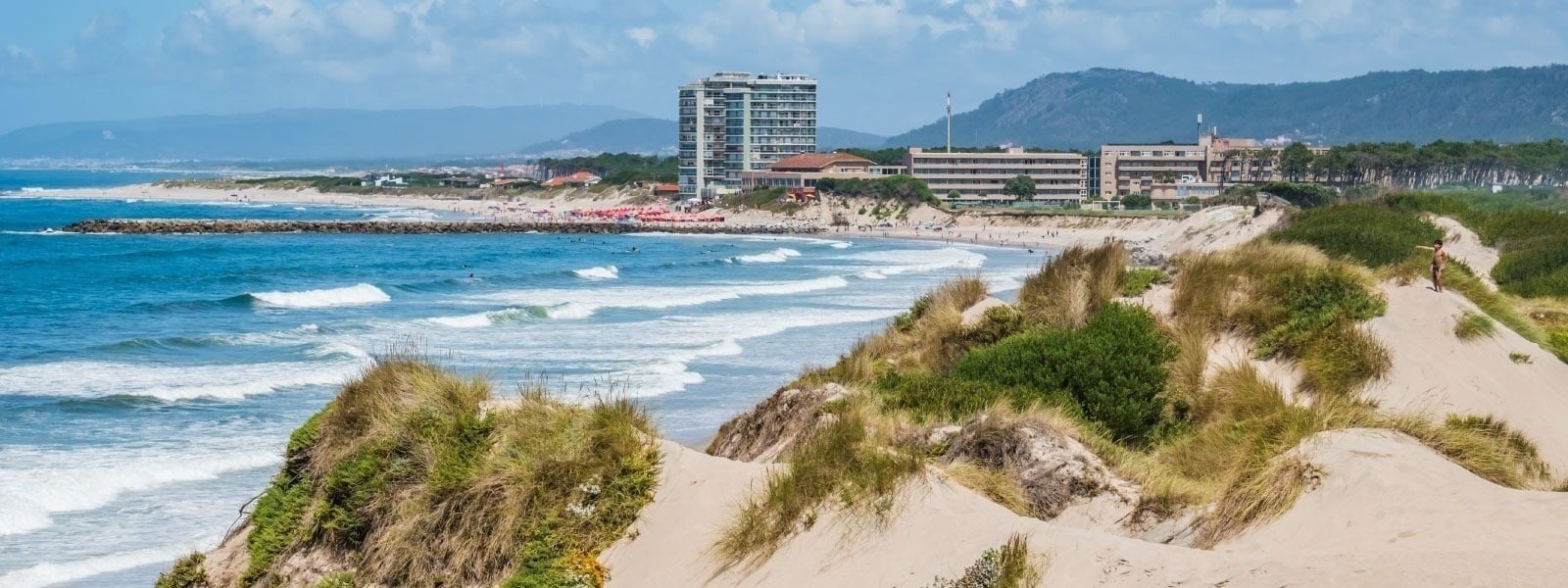 Une plage côtière avec des vagues, des dunes de sable verdoyantes et des bâtiments en arrière-plan sous un ciel nuageux.