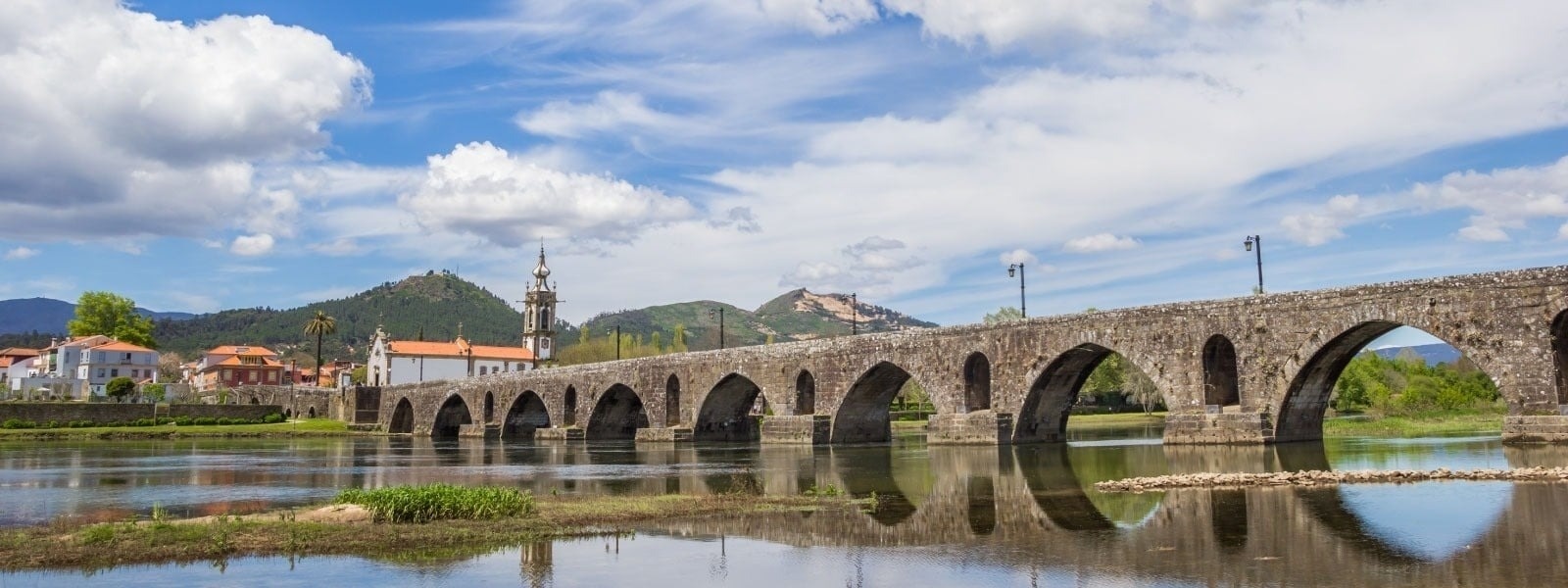 Uma ponte de pedra com arcos atravessa um rio, com uma vila e uma torre de igreja à esquerda e montanhas ao fundo, tudo sob um céu azul com nuvens.