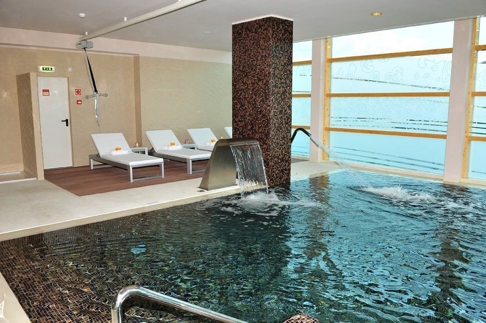 A imagem mostra uma moderna área de piscina interior com espreguiçadeiras, recursos de água, uma coluna decorativa e grandes janelas que proporcionam iluminação natural.
