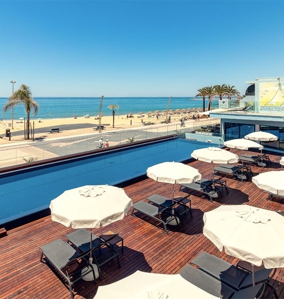 a swimming pool surrounded by chairs and umbrellas with a beach in the background
