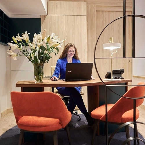 Oficina moderna con mujer en escritorio, flores y sillas naranja. U-Sense Sevilla Centro