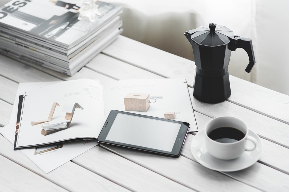 a stack of magazines sits on a table next to a cup of coffee and a tablet