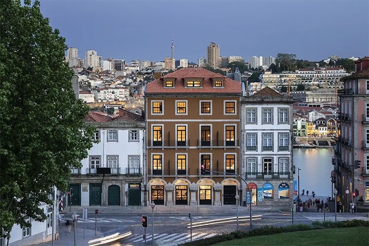 Uma piscina iluminada por luzes noturnas e rodeada por palmeiras sob um céu crepuscular com a lua visível.