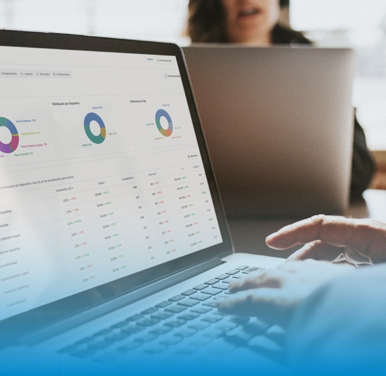 Manos tecleando en un portátil que muestra un panel de control con gráficos y tablas de datos analíticos, con otra persona trabajando en segundo plano.