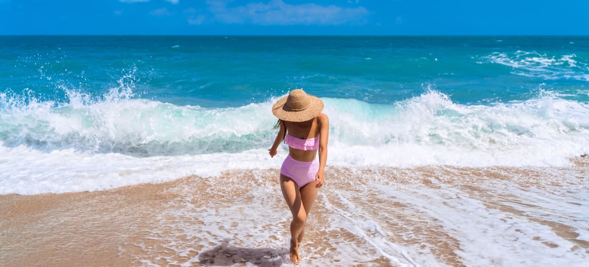 una mujer con traje de baño rosa y sombrero de paja camina por la playa