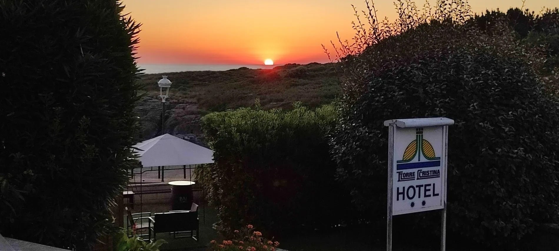Un vibrante atardecer sobre el mar ilumina un paisaje costero cubierto de vegetación con un cartel del "Torre Cristina Hotel" a la derecha.