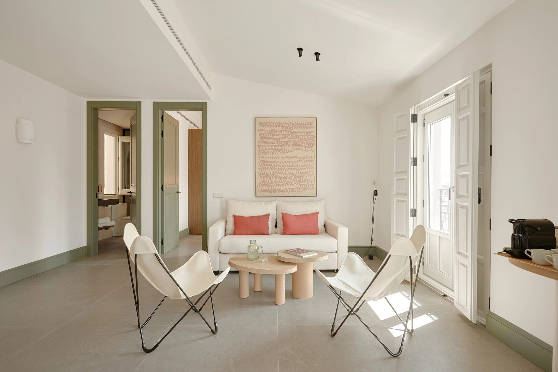 Luminosa sala de estar de diseño moderno con sofá claro, sillas mariposa blancas y un balcón de hierro forjado con vistas a edificios históricos. U-Sense Granada Gran Vía