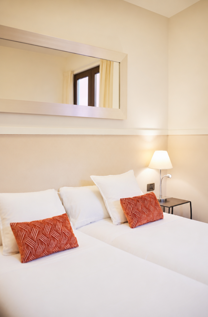 Dormitorio luminoso con camas gemelas en tonos neutros, cojines naranjas con estampado, espejo alargado sobre la pared y balcón con cortinas claras. U-Sense Sevilla Centro