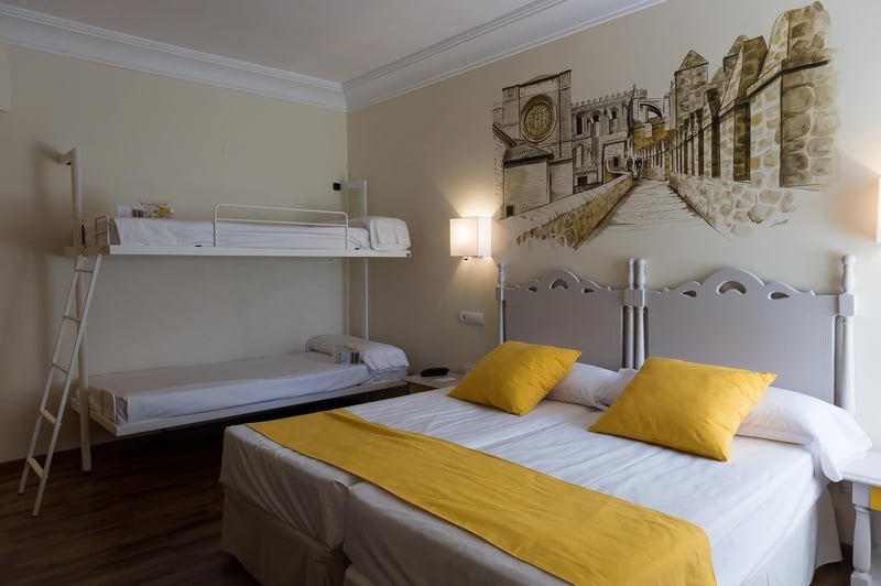 Una habitación de hotel con una litera y una cama grande con cabecero blanco, ropa de cama blanca con detalles amarillos, y un mural de una ciudad antigua en la pared.