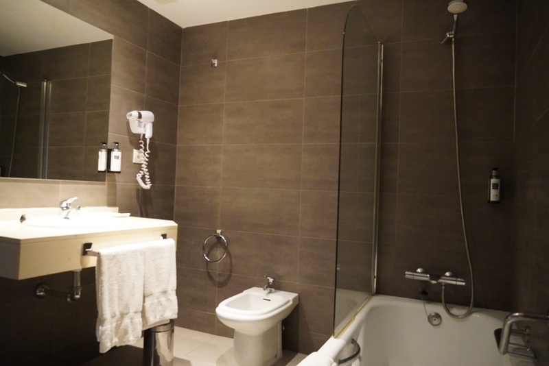 Un baño moderno con paredes de azulejos marrones, lavabo con espejo, secador de pelo, bidé y bañera con ducha.