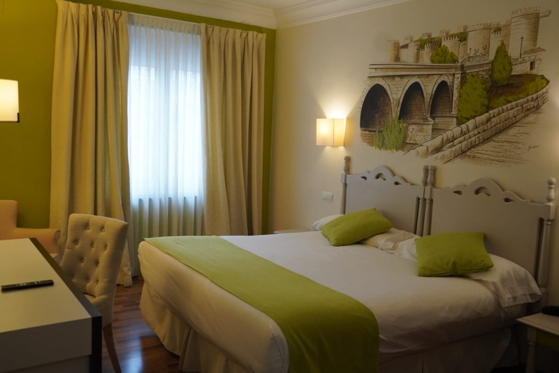 Habitación de hotel luminosa con una cama grande adornada con detalles verdes, un mural de un puente y un castillo sobre el cabecero y una ventana con cortinas claras.