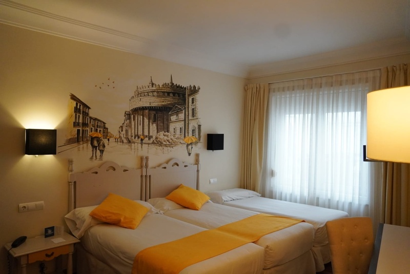 Una habitación de hotel luminosa presenta tres camas, dos de ellas adornadas con ropa de cama amarilla vibrante, frente a un mural que representa una calle urbana lluviosa con figuras sosteniendo paraguas y un edificio circular prominente.