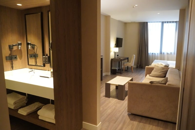 La imagen muestra una moderna habitación de hotel o apartamento que combina un área de tocador con lavabo y secadores de pelo a la izquierda, y un salón-dormitorio con un sofá, mesa de centro, escritorio, televisor y una cama junto a una ventana a la derecha.