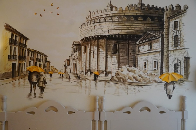 Un mural de pared en tonos sepia y amarillo retrata una calle lluviosa con edificios históricos y personas con paraguas, con un cabecero de cama blanco en primer plano.