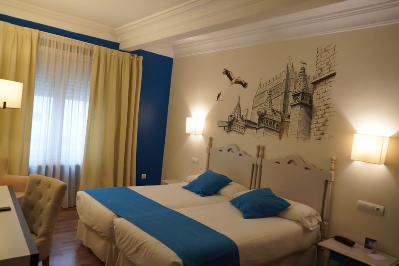 Una habitación de hotel con dos camas, cabeceros claros, detalles azules y un mural de un edificio histórico con cigüeñas en la pared principal.