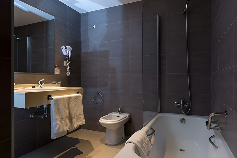 Un baño moderno y minimalista con azulejos grises oscuros, que incluye un lavabo con espejo, un bidé y una bañera con mampara de cristal.