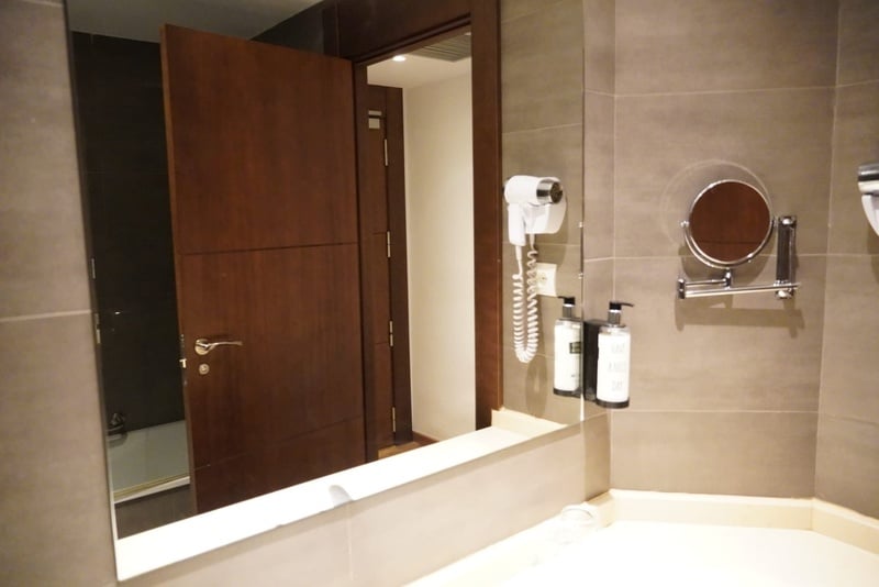 Un espejo de baño refleja una puerta de madera semiabierta, con un secador de pelo, dispensadores y un espejo de aumento visibles a la derecha.
