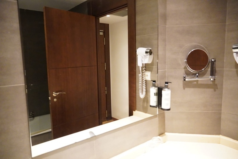 Un baño moderno presenta un espejo que refleja una puerta de madera entreabierta, junto a un secador de pelo, dispensadores de jabón y un espejo de aumento montados en la pared de azulejos grises.