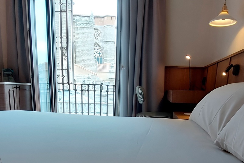 Una habitación de hotel moderna presenta una cama blanca y una vista a una majestuosa catedral de piedra a través de su balcón.