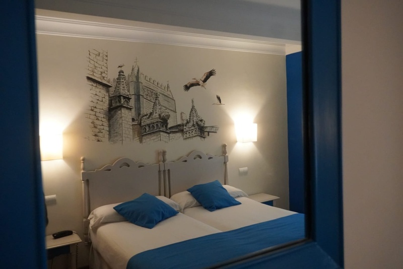 Una habitación de hotel con dos camas individuales, ropa de cama azul y un mural de una catedral con cigüeñas volando se observa a través de un marco azul.