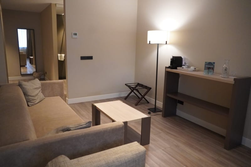 Una moderna y acogedora sala de estar, probablemente de un hotel, con un sofá, una mesa de centro, una lámpara de pie iluminada, una estación de café y un espejo que refleja un pasillo al fondo.
