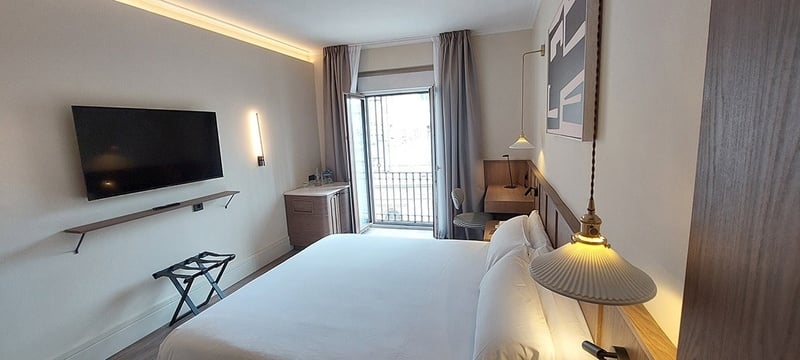 Una habitación de hotel moderna y luminosa con una cama grande, televisión montada en la pared, un pequeño escritorio y un balcón.