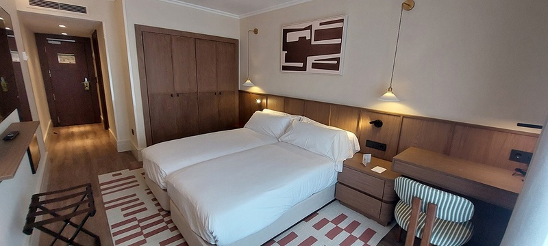 Habitación de hotel luminosa con dos camas individuales juntas, mobiliario de madera, una alfombra de patrones geométricos y decoración moderna.