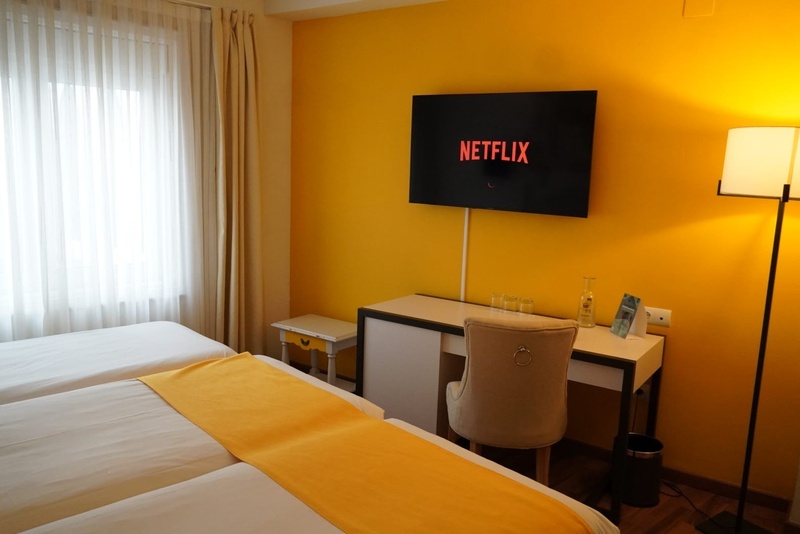 es: Una vibrante habitación de hotel cuenta con una pared amarilla brillante, un televisor mostrando Netflix, dos camas individuales, un escritorio con silla y una lámpara de pie, con una ventana y cortinas claras a la izquierda.