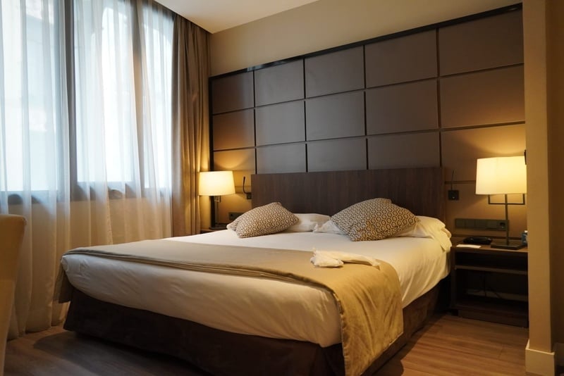 Un moderno dormitorio de hotel se ve con una cama grande, cabecero de paneles oscuros, lámparas con luz cálida y una ventana con cortinas claras.