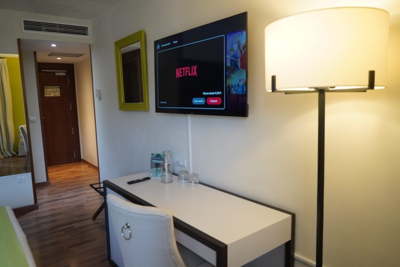 La imagen muestra el interior de una habitación de hotel con un televisor que exhibe Netflix, un espejo enmarcado en verde, un escritorio con silla y una lámpara de pie, con un pasillo y una puerta de madera al fondo.