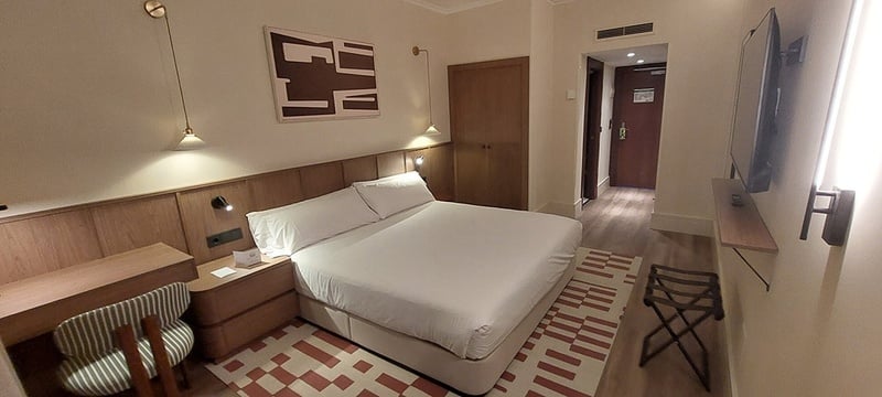 Una moderna y acogedora habitación de hotel cuenta con una cama doble con ropa de cama blanca, mobiliario de madera clara, una alfombra con diseño geométrico y una televisión montada en la pared.
