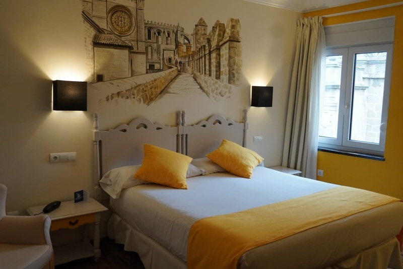 Una habitación de hotel con una cama vestida de blanco y detalles amarillos, un mural arquitectónico detrás del cabecero y dos lámparas de pared.
