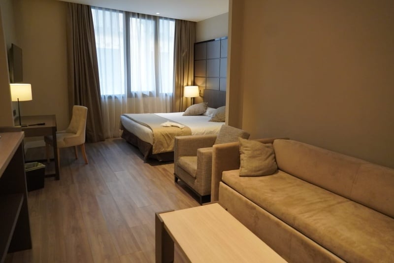 La imagen muestra el interior de una habitación de hotel con una cama doble, un escritorio con silla, un sofá y un sillón, todo en tonos neutros y con luz natural entrando por la ventana.