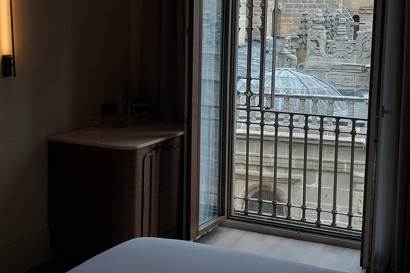 El interior oscuro de una habitación contrasta con la vista de un edificio de piedra histórico y detallado a través de una ventana abierta con barandilla.