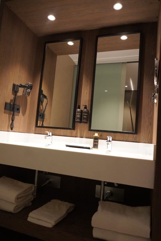 Un moderno tocador de baño doble con lavabos, espejos, un secador de pelo montado en la pared, artículos de tocador y toallas, todo en un entorno con paneles de madera.