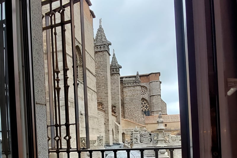 Desde una ventana enrejada, se observa una antigua catedral de piedra con intrincados detalles arquitectónicos bajo un cielo nublado.