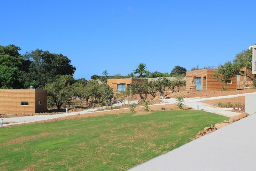Varias cabañas modernas de madera se encuentran dispersas en una ladera con césped verde y árboles bajo un cielo azul claro.