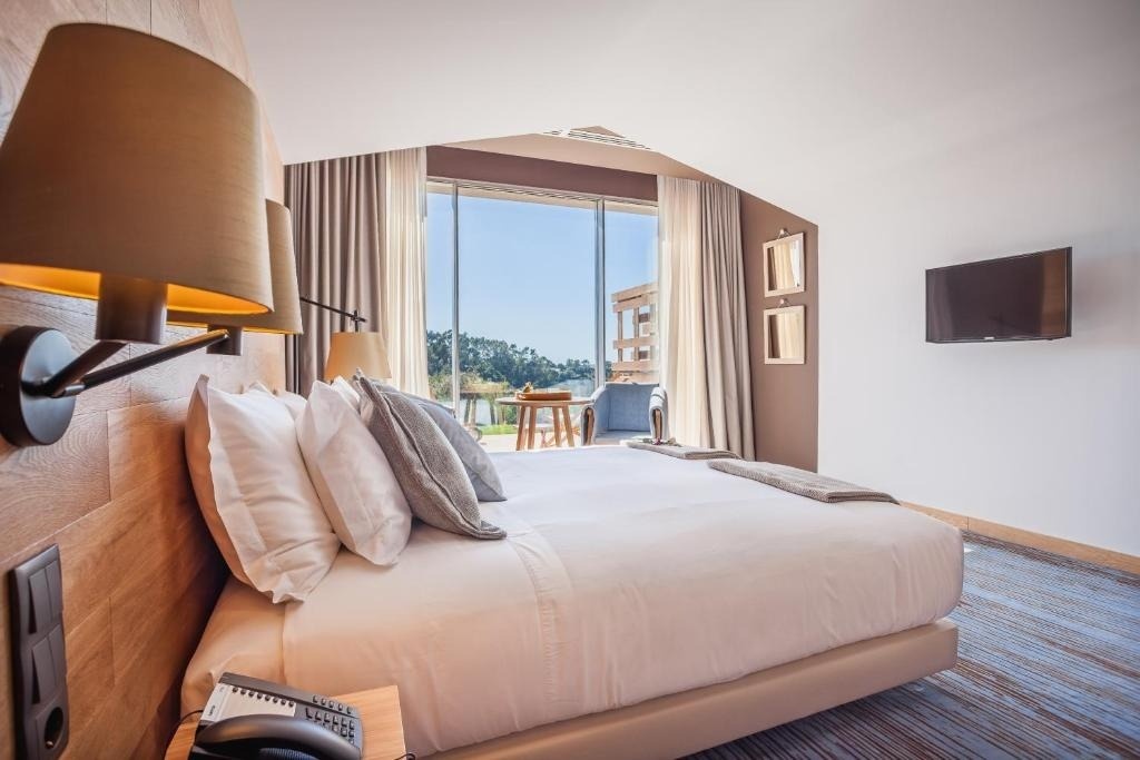 Una habitación de hotel moderna con una cama grande, lámparas de pared, un televisor y una ventana amplia que da a un balcón con vistas a la naturaleza.