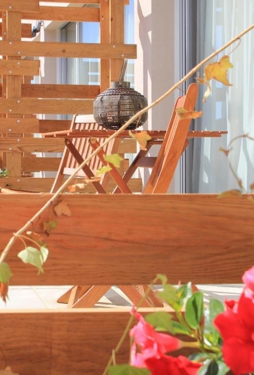 Un balcón soleado presenta muebles de madera, una linterna decorativa, y vegetación con flores rojas en primer plano.