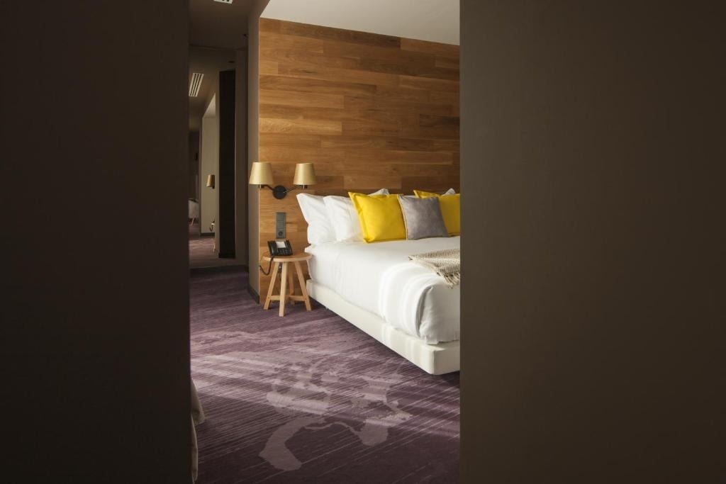 Se observa una moderna habitación de hotel con una cama, una pared de madera, almohadas vibrantes y una distintiva alfombra púrpura.