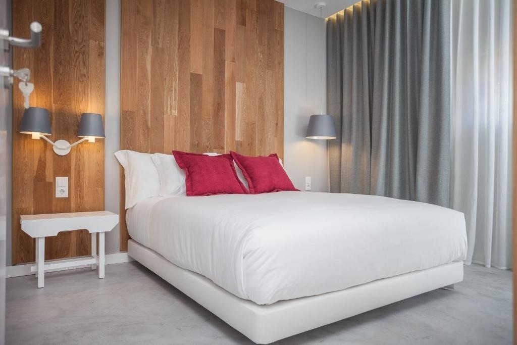 Un dormitorio moderno y luminoso presenta una cama con ropa blanca y almohadas rojas, un cabecero de madera y cortinas grises.