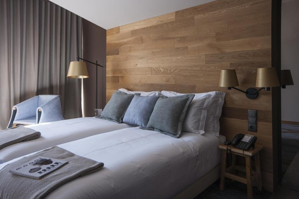 Una habitación de hotel moderna y acogedora con dos camas individuales juntas, un cabecero de madera clara, cortinas grises y almohadas en tonos azules y grises.