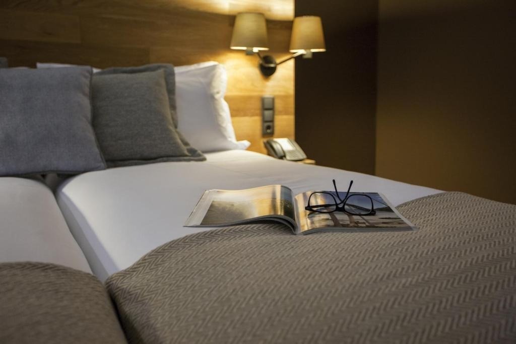 Una cama de hotel con un libro abierto y gafas de lectura sobre una manta, iluminada cálidamente por lámparas de pared.
