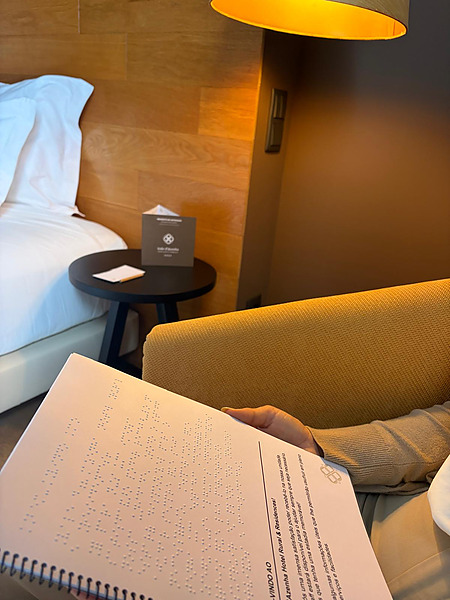 Guía en Braille abierta en la habitación, con la cama y la mesa auxiliar al fondo.