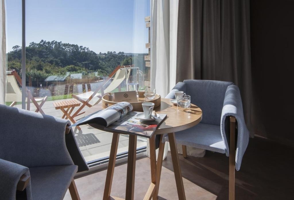 Un interior moderno y luminoso presenta una mesa de madera con tazas de café, una revista y una jarra de agua, junto a dos sillas azules claras, con un frondoso paisaje natural visible a través de un gran ventanal.