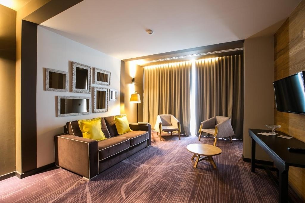 Esta es una sala de estar moderna y bien iluminada, con un sofá marrón con cojines amarillos, sillones grises, una alfombra estampada y una pared decorada con espejos y una pantalla de TV.