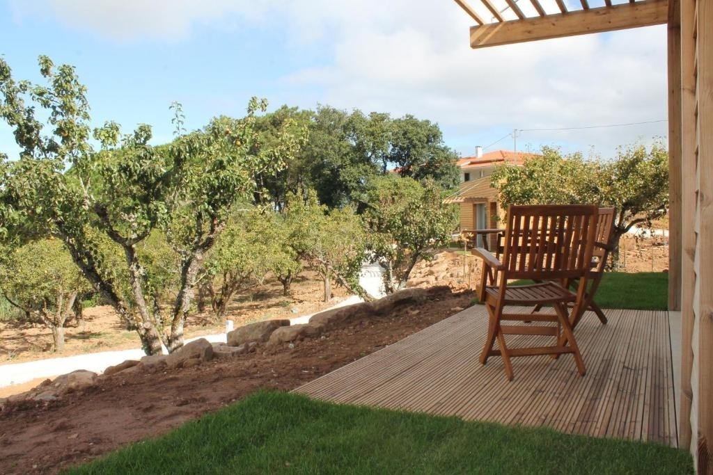 es: Desde una terraza de madera con sillas, se aprecia un huerto frutal que desciende hacia una casita, bajo un cielo azul con nubes.