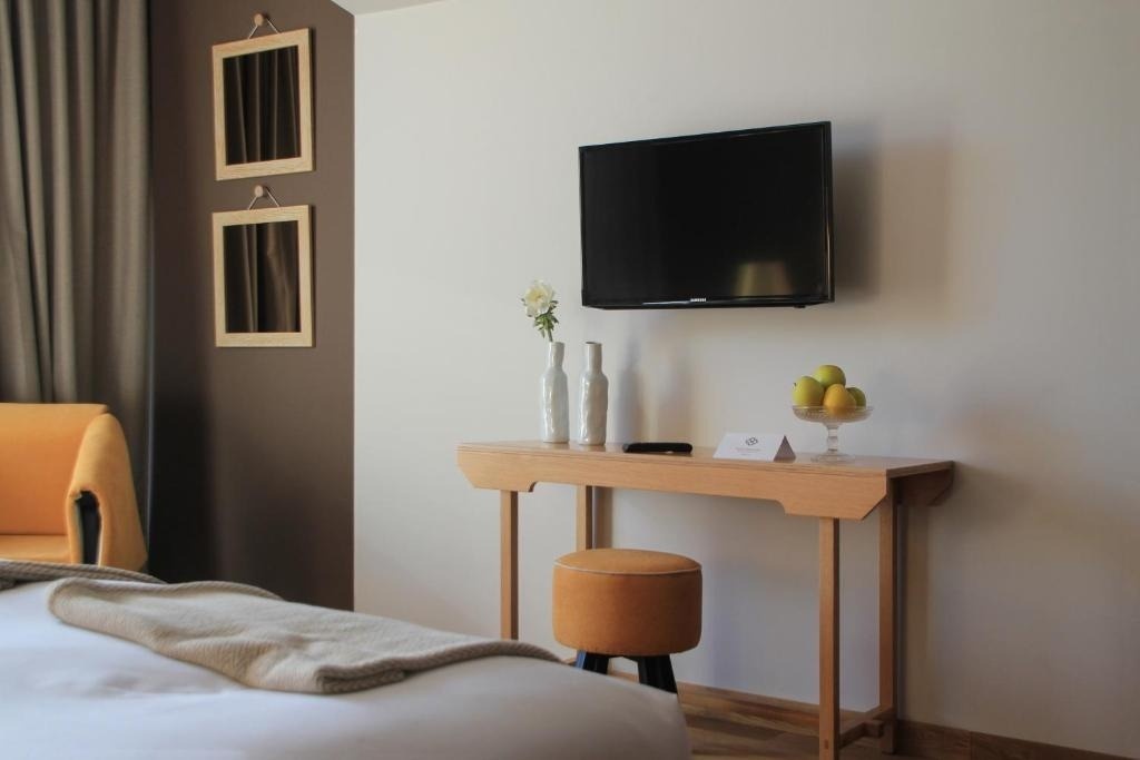 Una habitación moderna presenta una cama con una manta, un sillón naranja, un televisor montado sobre una mesa de madera con jarrones y un frutero, y dos espejos decorativos en la pared.