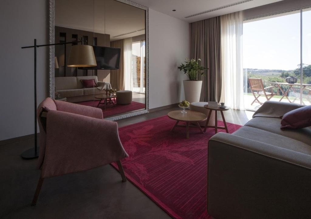 Un moderno salón luminoso está decorado con un gran espejo, una alfombra de color magenta y un ventanal con vistas a una terraza exterior.