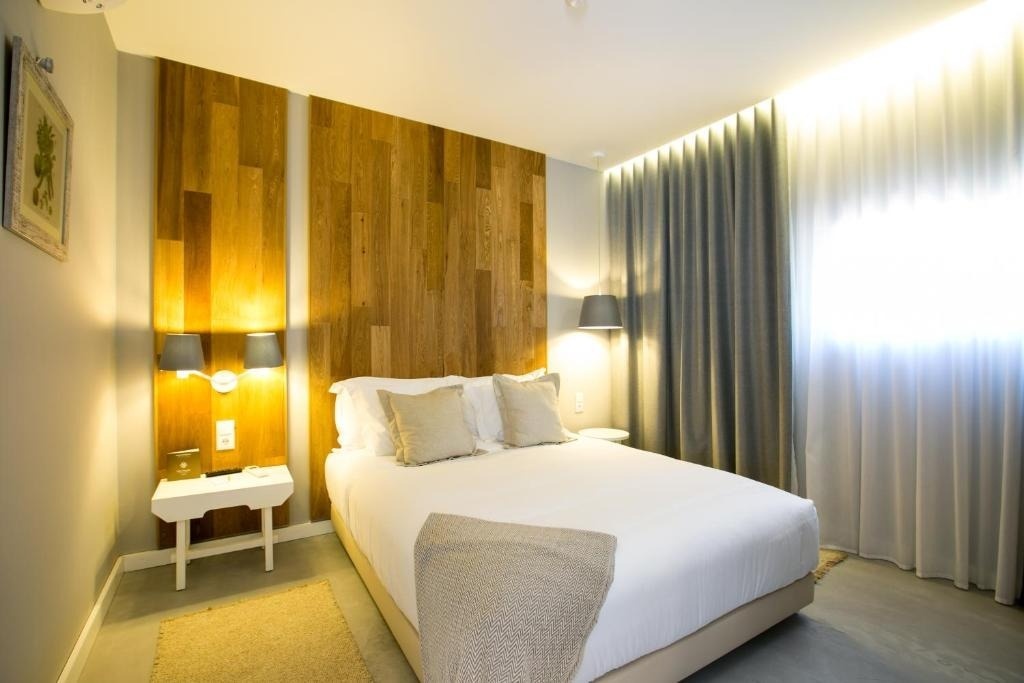 Un moderno dormitorio presenta una cama con cabecero de madera, lámparas de pared y cortinas de tela en una ventana luminosa.